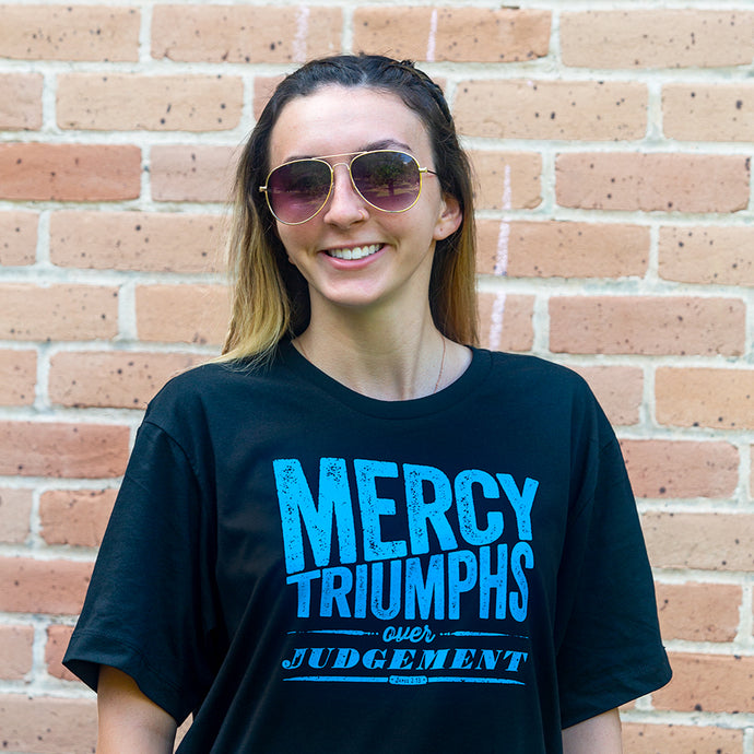 Brunette model with sunglasses wearing Mercy triumphs over judgement, James 2:13,  with textured dynamic design printed in bright blue on black Christian t-shirt. Shot outdoors with light brick background.