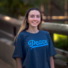 Load image into Gallery viewer, Brunette model wearing a Navy blue Christian t-shirt with &quot;Peace be with you&quot;  in light blue on the front. Photographed outdoors in a park at dusk with bokeh.