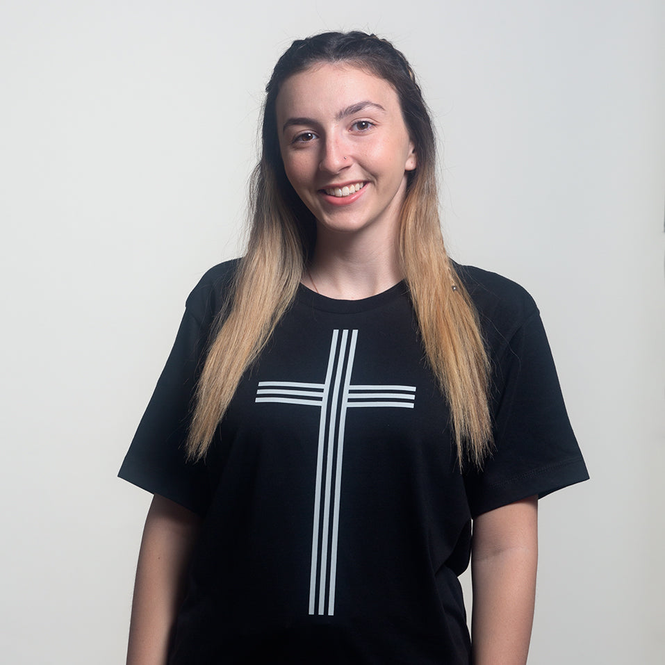 Model wearing black t-shirt with simple Christian cross consisting of 3 vertical lines and 3 horizontal lines printed in white. Shot in studio on white background.