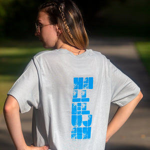 Woman photographed outdoors with "Hallelujah God be Praised" printed in light blue on back of light gray t-shirt.