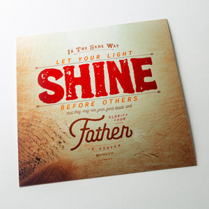 Angled shot of Metallic photo print with Matthew 5:16 in textured type with wooden background