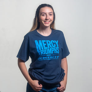 Brunette model wearing "Mercy triumphs over judgement," James 2:13,  with textured bold design printed in iight blue on navy blue Christian t-shirt. Shot in studio on light gray backgroud.